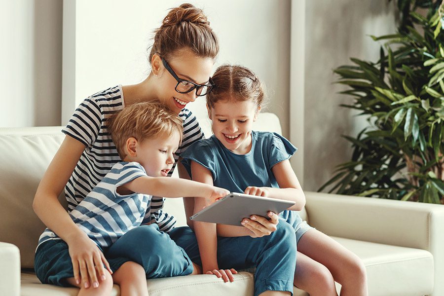 Client Center - A Mother and Her Two Children are Sitting Together on a Sofa Using a Tablet to Video Chat at Home