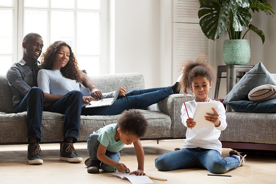 Personal Insurance - A Father and Mother are Sitting Together on a Sofa Using a Laptop While Their Two Children are Drawing Pictures in Their Books on the Floor at Home
