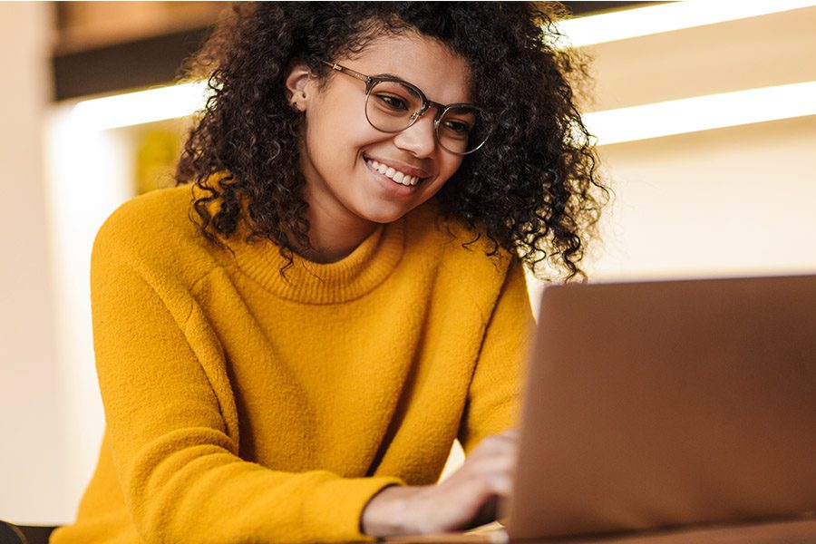 Blog - Young Woman in Yellow Sweater Smiling and Looking at Her Laptop While Sitting at a Desk