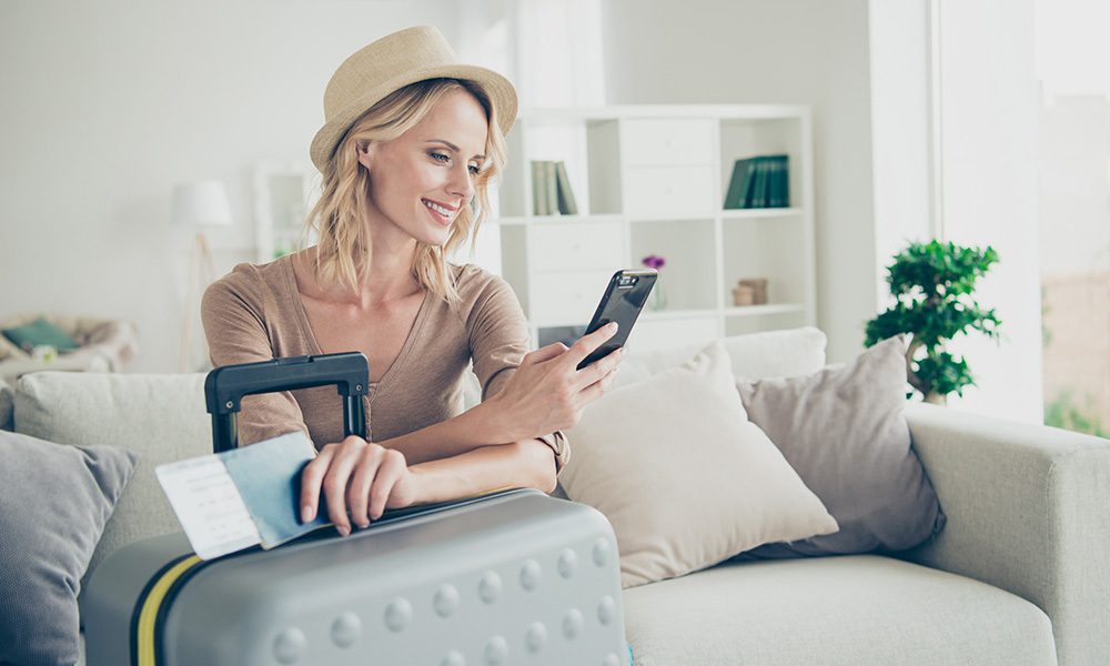 Blog Post - Happy Woman Looks at Her Phone at Home as She Prepares for a Trip
